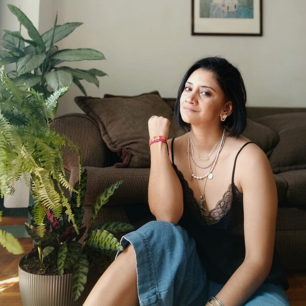 Un retrato profesional y cercano de Maguie, la tarotista de Maguie Tarot en Venezuela, sentada en un ambiente cálido y sereno con plantas naturales de fondo. Ella viste una blusa negra de encaje y jeans, proyectando una presencia auténtica, consciente y empática ideal para el acompañamiento en sesiones de tarot terapéutico y evolutivo.
